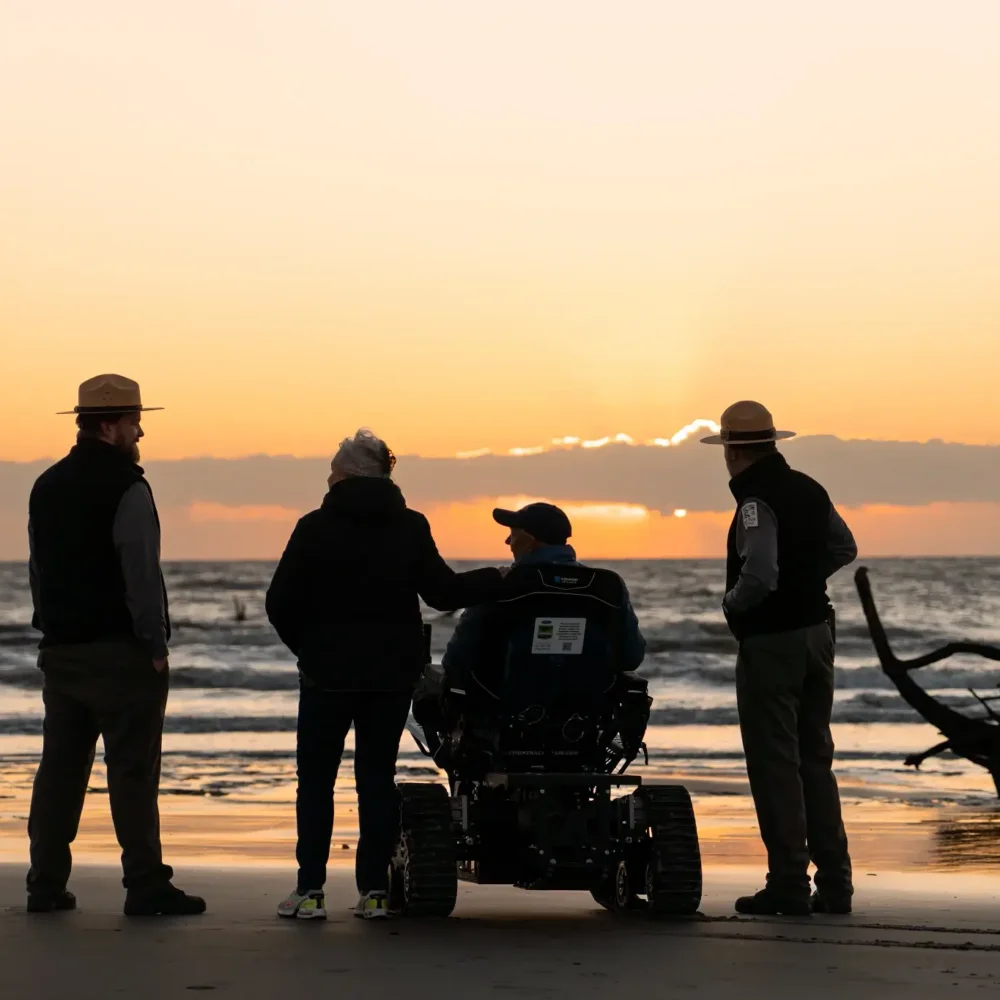 A man in the Action Trackchair AXIS with a female and two park rangers on the beach at sunset